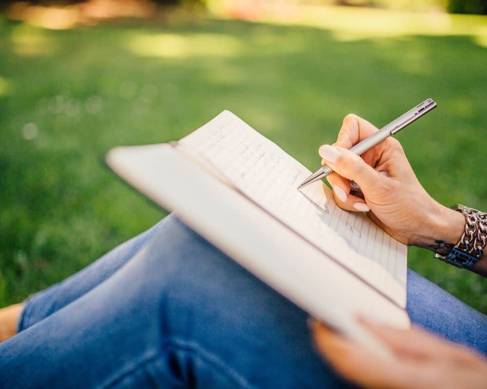 Image of a woman sitting outside, writing in a journal. Concept of prayer journaling.