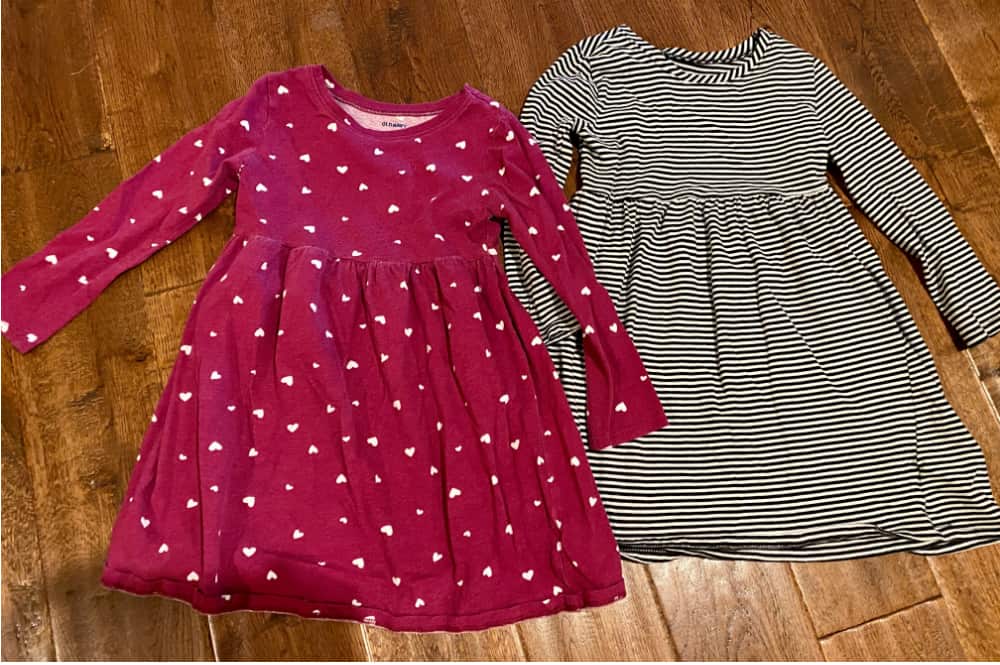 Image of a pink toddler dress with hearts and a black-and-white striped toddler dress side by side on a wooden floor. Concept of selling clothing lots versus single items on Marketplace.