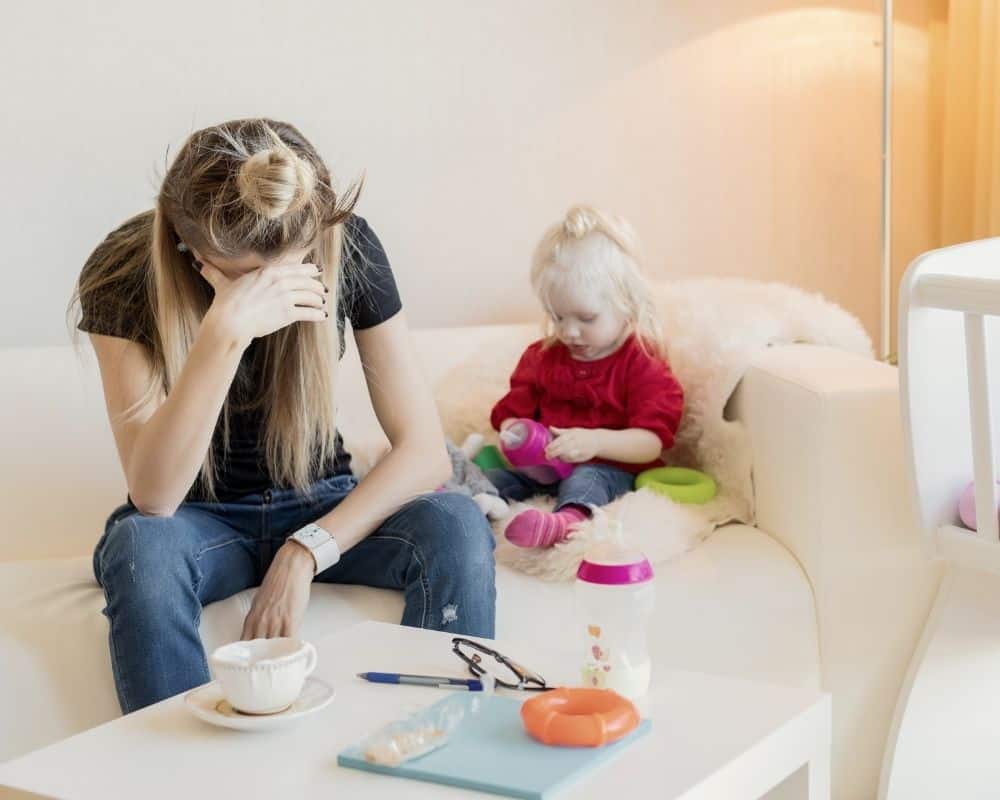 Image of struggling mom sitting on a couch with her head in her hands while her toddler plays in the corner.