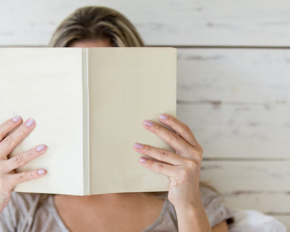 woman holding up a book in front of her face, standing against a wooden background.