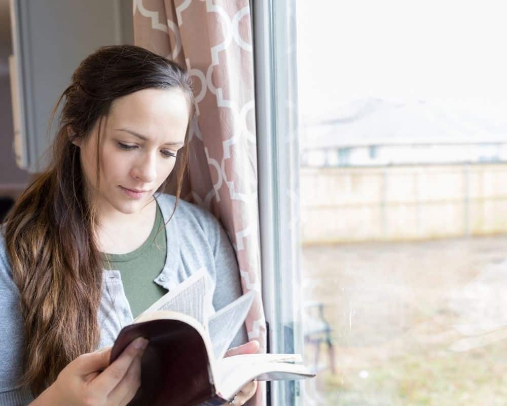 Image of a young woman standing by a window, holding a Bible and studying it. Concept of understanding the basis of Christian faith.
