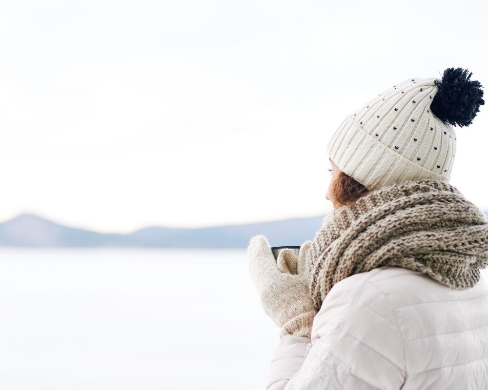 Woman standing outside, bundled up in warm clothing and sipping coffee, looking contemplative.