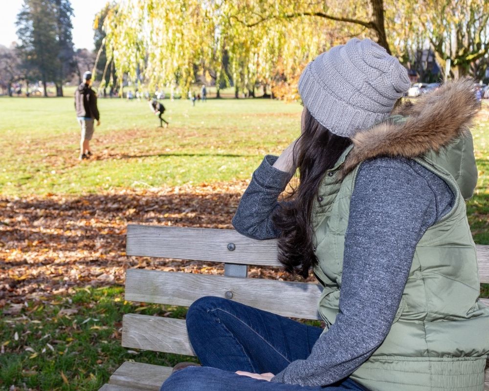 Woman sitting on a park bench observing her family playing, from a distance.