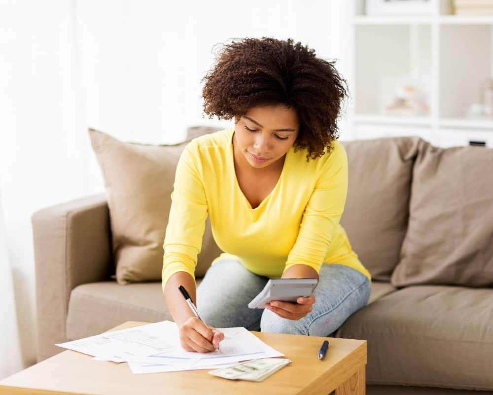 Woman sitting on a couch with a calculator and pen in hand, writing a monthly budget plan.