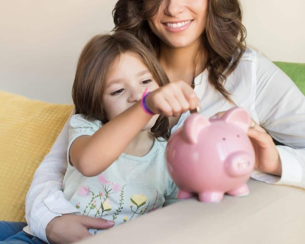 Little girl placing coins into a piggy bank, sitting with her mom on a couch.
