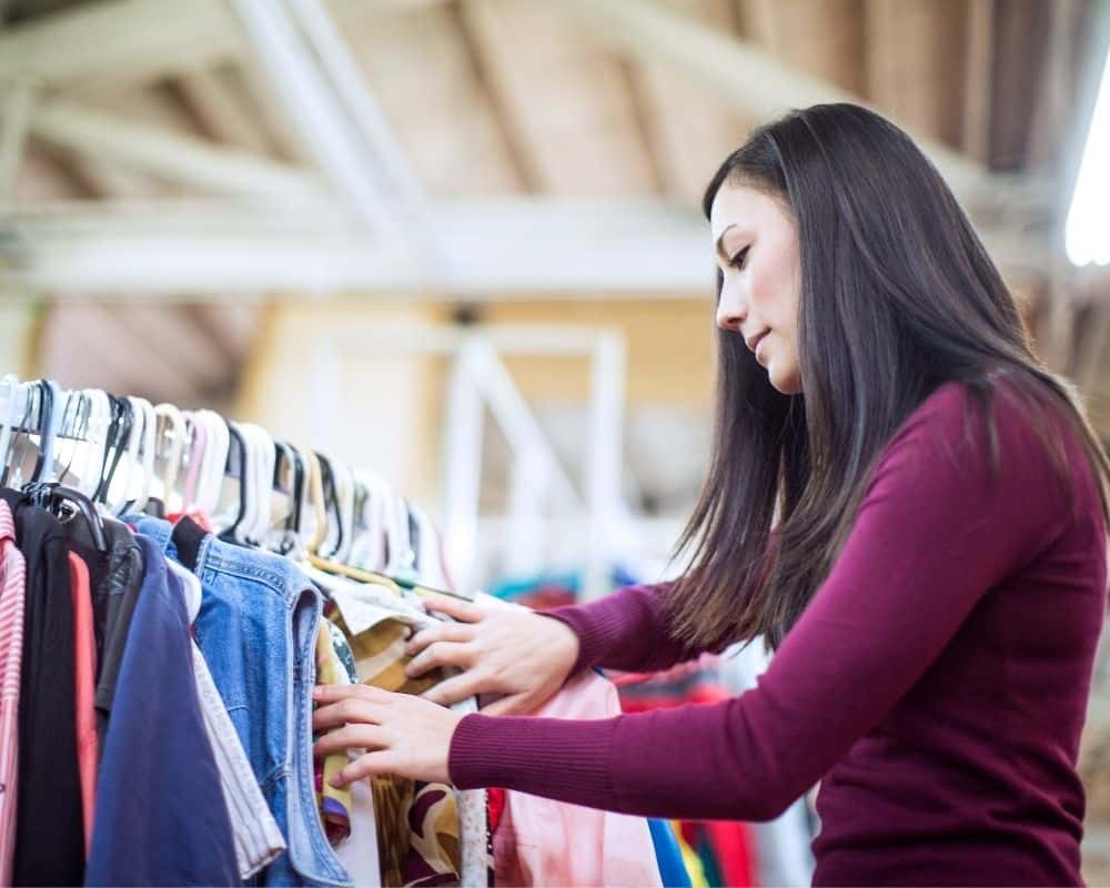 Woman browsing clothing racks at a thrift store- frugal living idea for families.