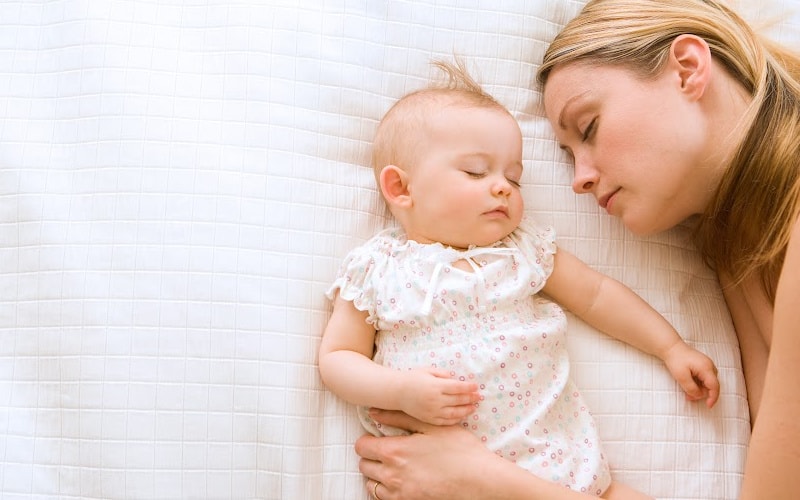 A mother sleeping beside her infant daughter on a white bedspread.