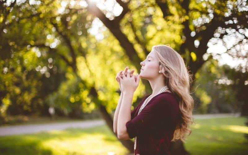 A blonde woman praying outside beneath a tree.