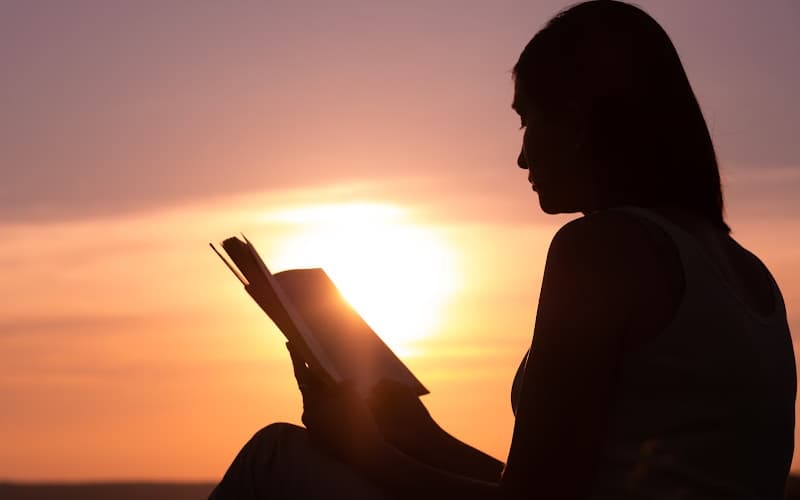 Woman sitting outside reading the Bible with the sunset in the background.