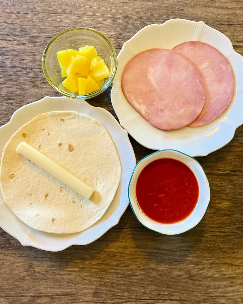 Hawaiian air fryer pizza roll ingredients on plates and dishes against a wooden background.