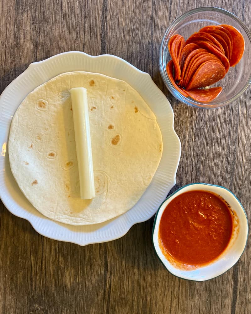 A flour tortilla on a plate with a mozarella stick in the middle, alongside a handful of pepperoni in a cup and another cup of marinara sauce.