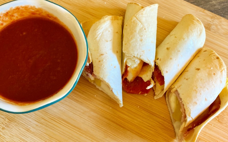 Finished air fryer recipe for kids: pizza roll ups- 4 slices on a cutting board with marinara dipping sauce pictured to the side.