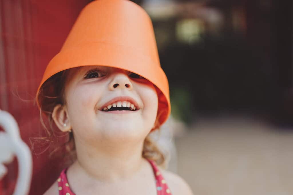 Little girl smiling with a flower pot on her head.