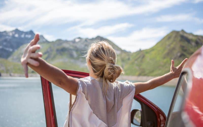 Woman standing outside her car with arms outstretched, facing a beautiful mountain range- concept of overcoming struggles.