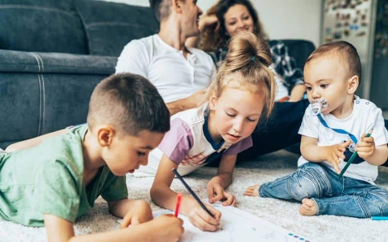 Two young kids doing an art project on the living room floor with a baby watching on, with parents on the couch in the background, smiling.