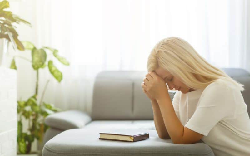 Woman knelt down on an ottoman in the living room praying with a Bible in front of her.