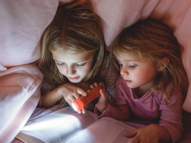 Two sisters under a blanket with a flashlight, reading a book together.