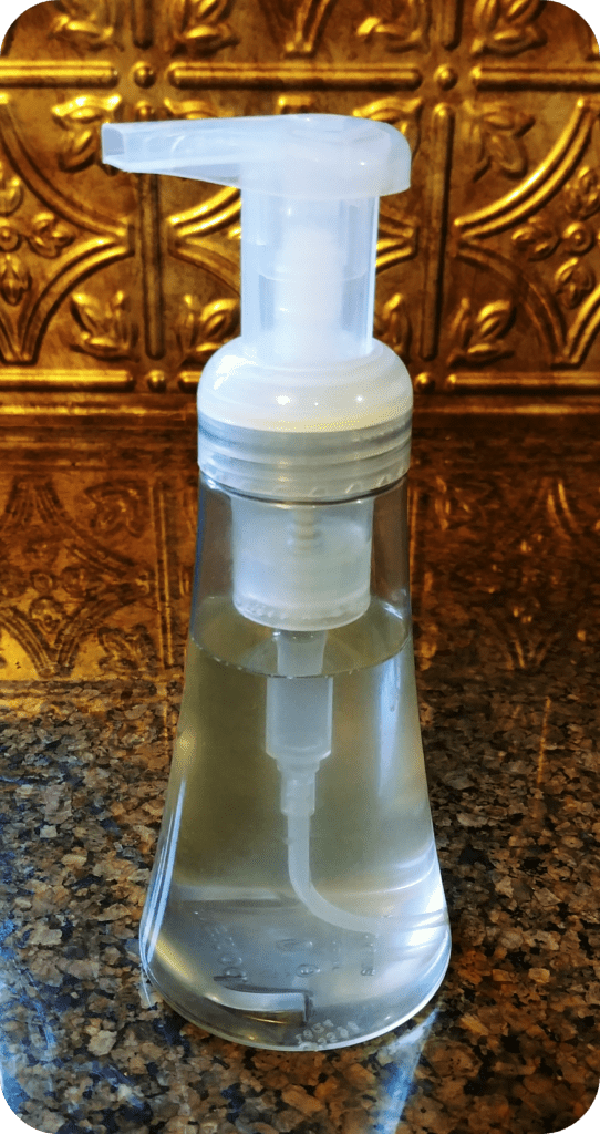 Homemade foaming hand soap in a plastic container sitting on a kitchen counter with a bronze tile background.