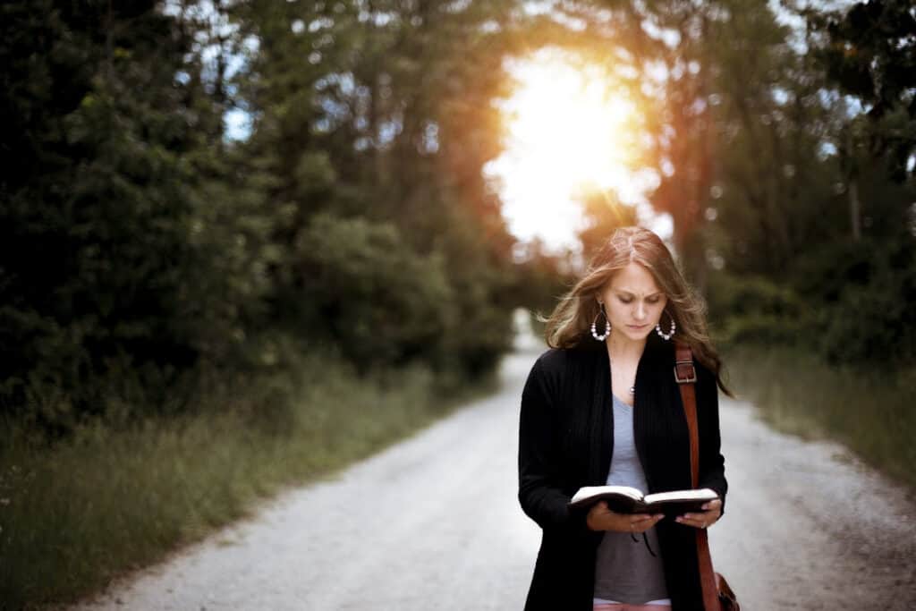 Woman standing on a dirt road reading the Bible with the sunset in the background.