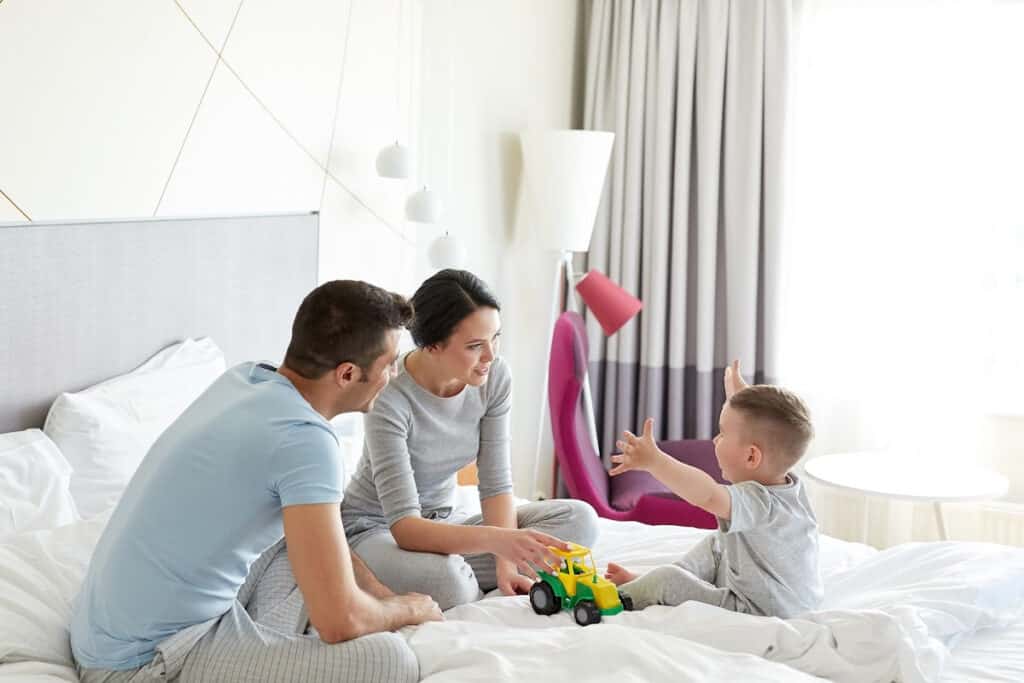 Mom and dad sitting in bed in toddler son's room, playing trucks with him before bedtime.