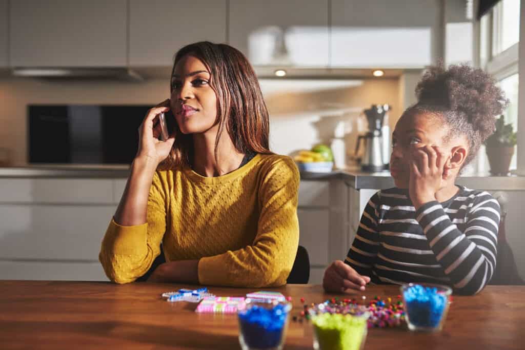 A distracted mom on her cell phone while her preteen daughter stares at her, making a craft and waiting on mom to pay attention to her.