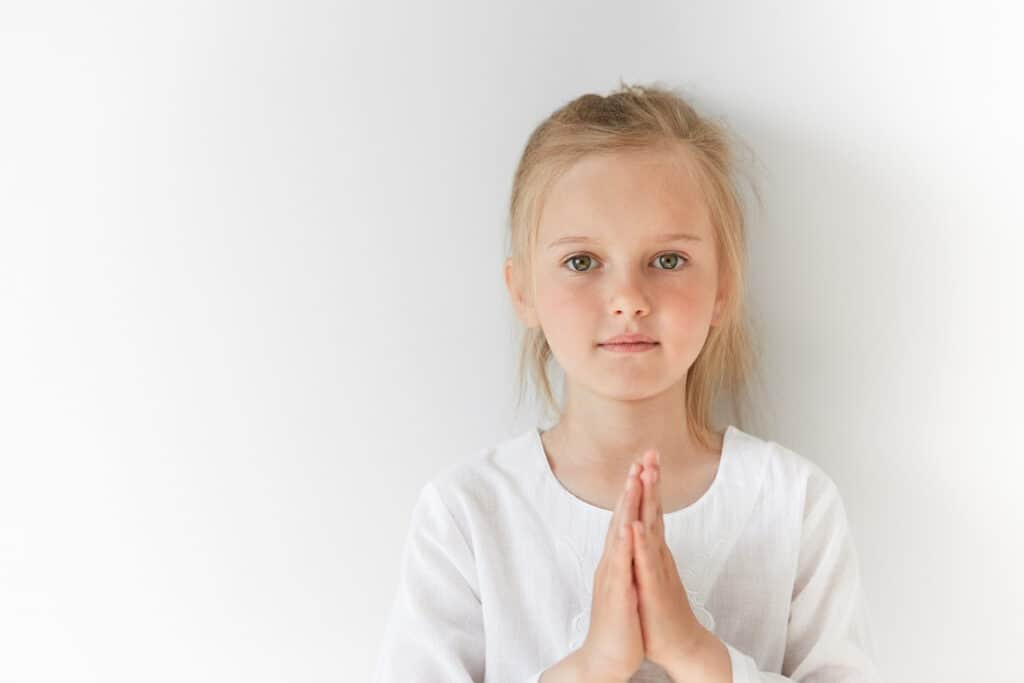 young blond girl with hands folded in prayer, standing in front of a white background.