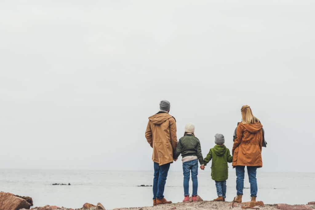 Young family hand in hand, staring out across a beach together on a cloudy day.