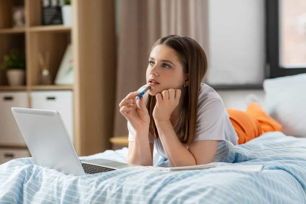 Teen girl sitting on her bed looking into space, thinking. With a laptop sitting on her bed and a notebook. Concept of having questions about faith.