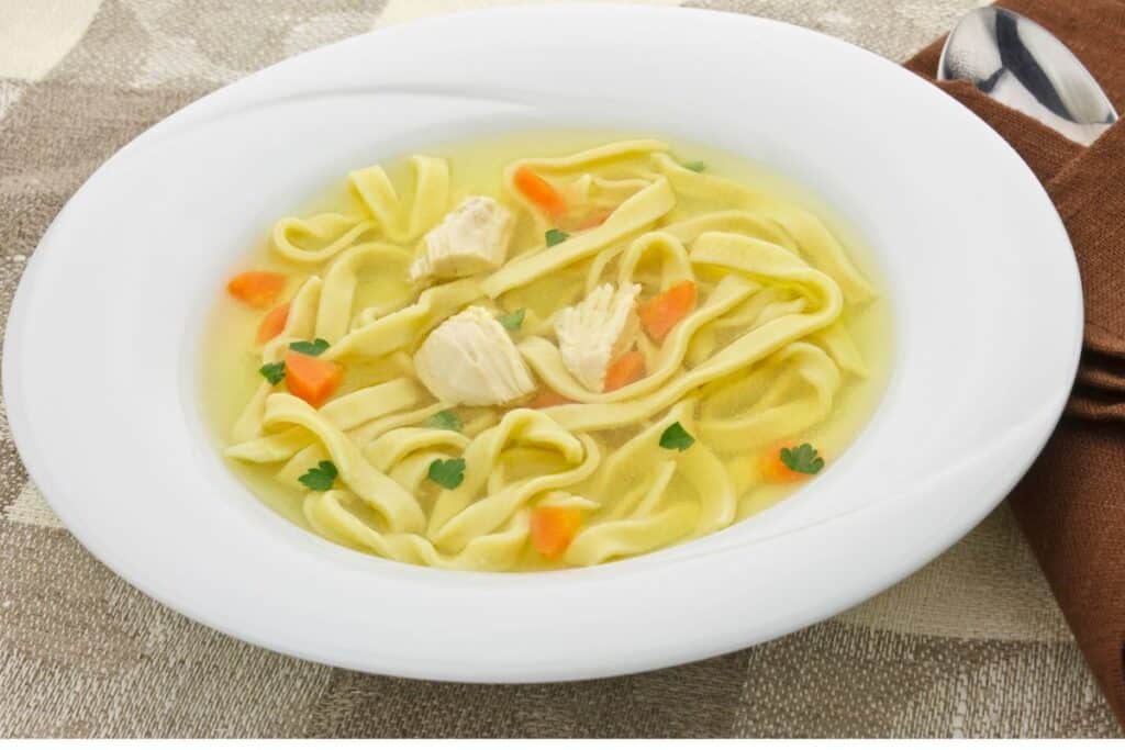 Bowl of chicken and noodle soup with vegetables on a tan checkered placemat.