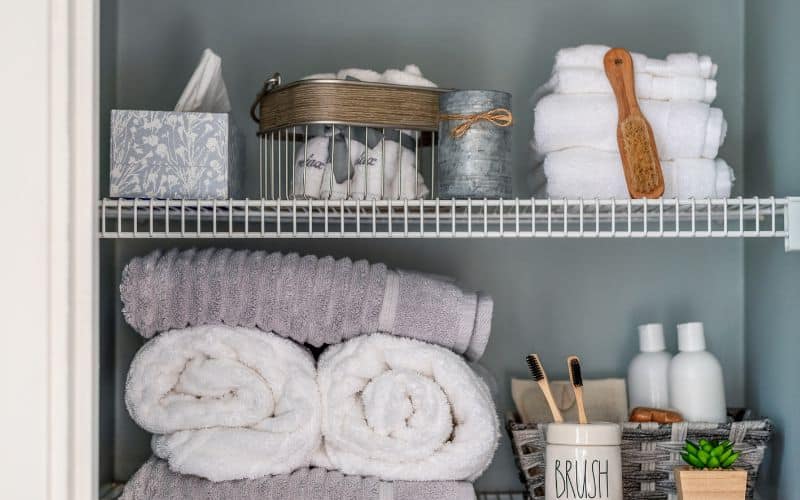 Organized bathroom linen closet with neatly rolled towels and toiletries in a grey woven basket.