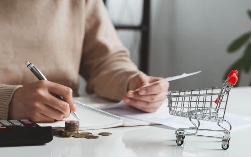 Woman calculating her grocery budget on paper with a calculator and tiny shopping cart sitting on her desk.