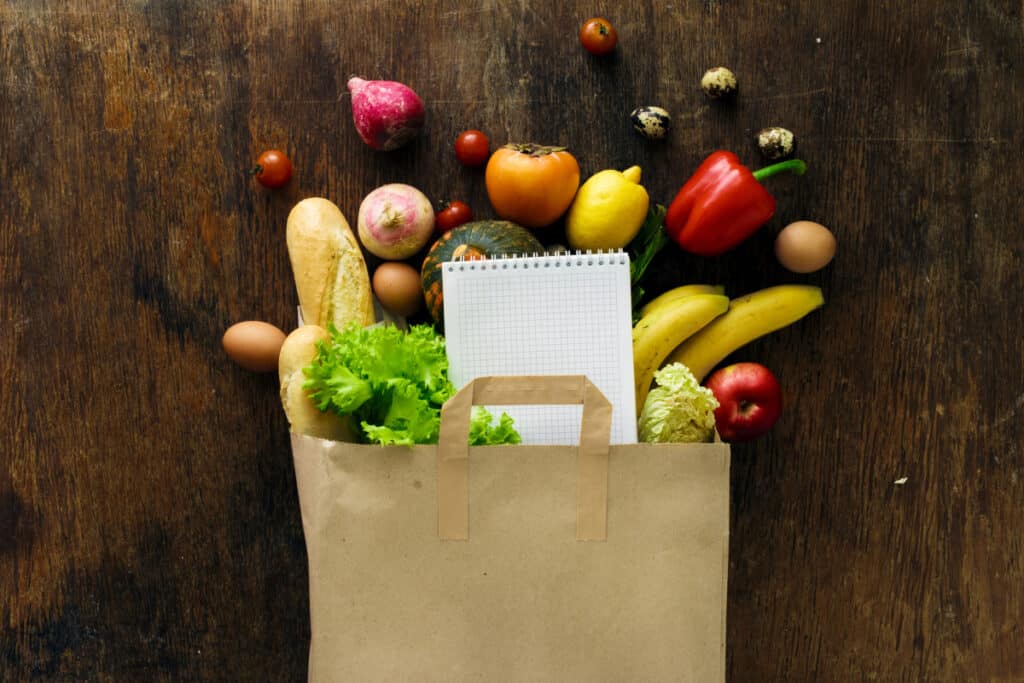 paper grocery bag with fresh produce and a notebook spilling out onto a wooden background