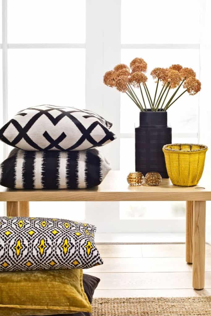 Storage bench with decorate pillows stacked on top and below with a blue vase and yellow basket beside it.