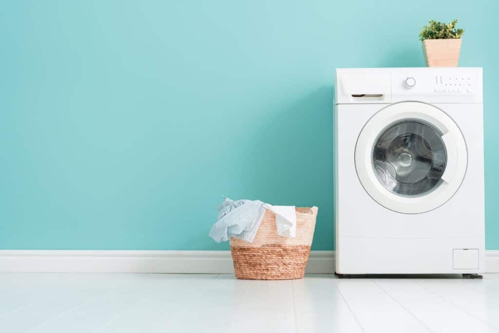 Front-load washing machine in front of a teal blue laundry room wall with a basket of dirty clothes on the floor.