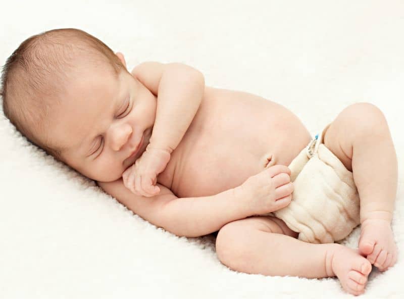 Newborn baby boy asleep on a fluffy white blanket, while wearing just a prefold cloth diaper with diaper pins.