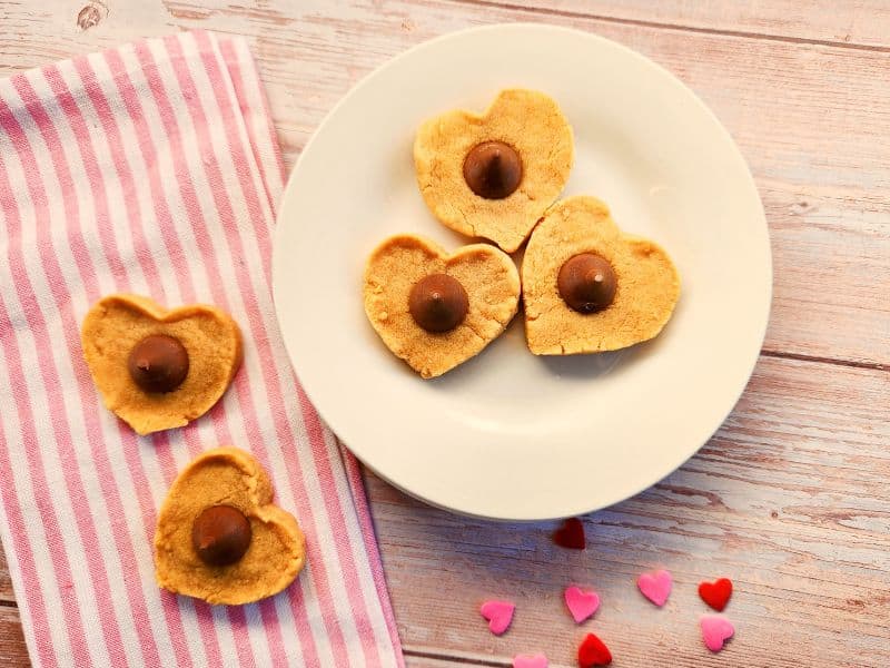 3 heart shaped peanut butter cookies on a plate on a whitewashed wood table with a pink striped tea towel beside it and two more cookies on top.