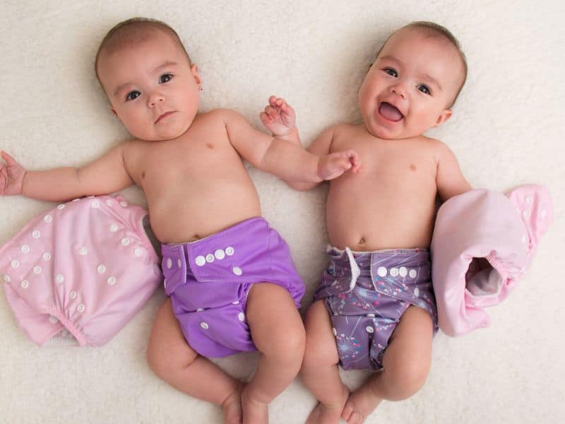 Twin babies lying on a fluffy white blanket, both wearing colorful cloth diapers and holding cloth diapers.