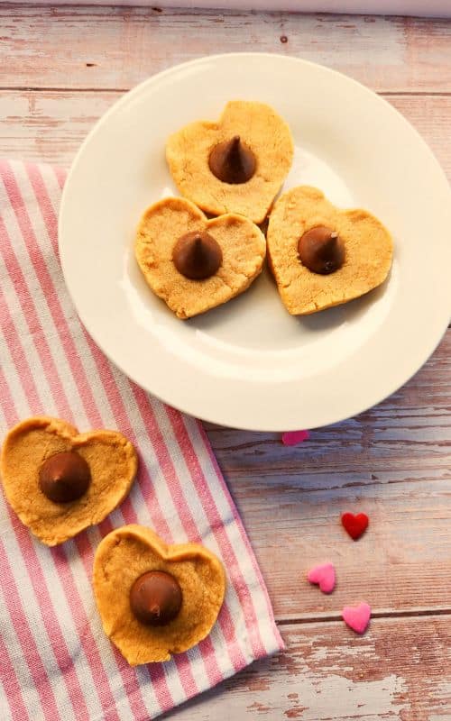 vertical photo of three heart-shaped cookies on a white plate with chocolate kisses in the middles; and two more cookies sitting on a pink and white striped tea towel below.