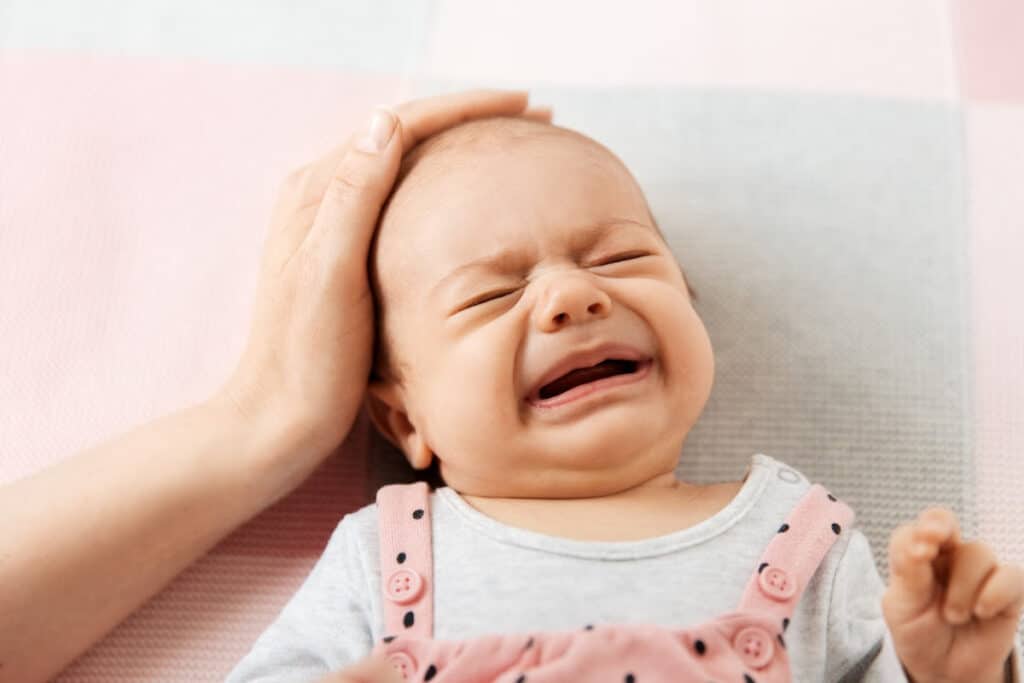 Crying newborn baby girl on a soft pink blanket with mom's hand cradling her head.