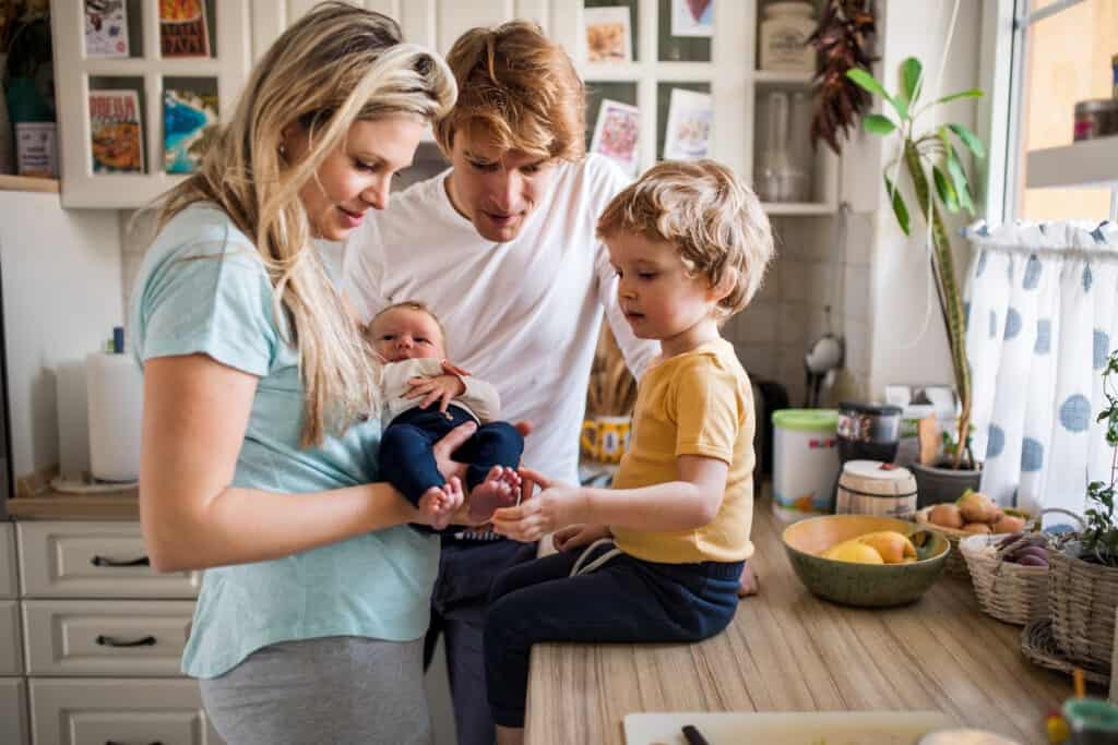 Mom and dad with a toddler in the kitchen, holding and staring at newborn sibling.