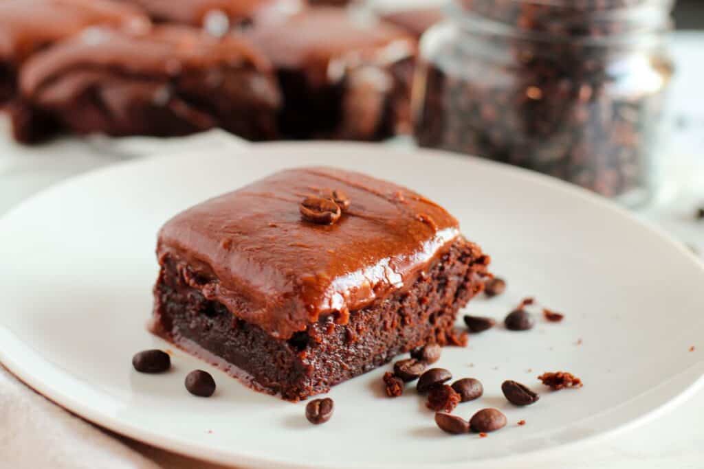 Chocolate frosted mocha brownies on a white plate with coffee beans scattered on plate and ingredients in background.