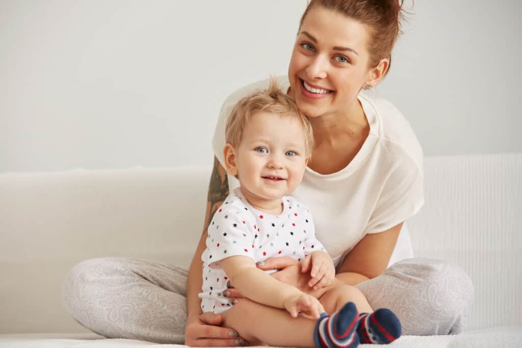 Mom with toddler boy smiling against a white couch and white wall.