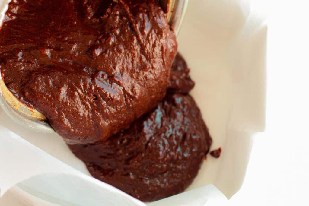 Brownie batter being poured from sauce pan into parchment-lined baking pan.