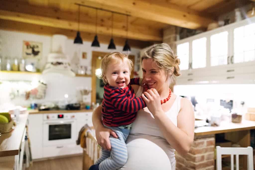 Pregnant young mom holding a toddler boy on her hip, smiling and laughing in the kitchen together.