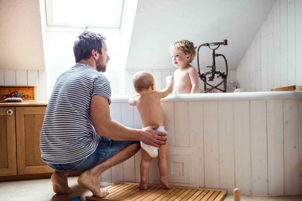 Dad bathing toddler in a large old-fashioned tub while holding a baby who is standing up against the outside of tub.