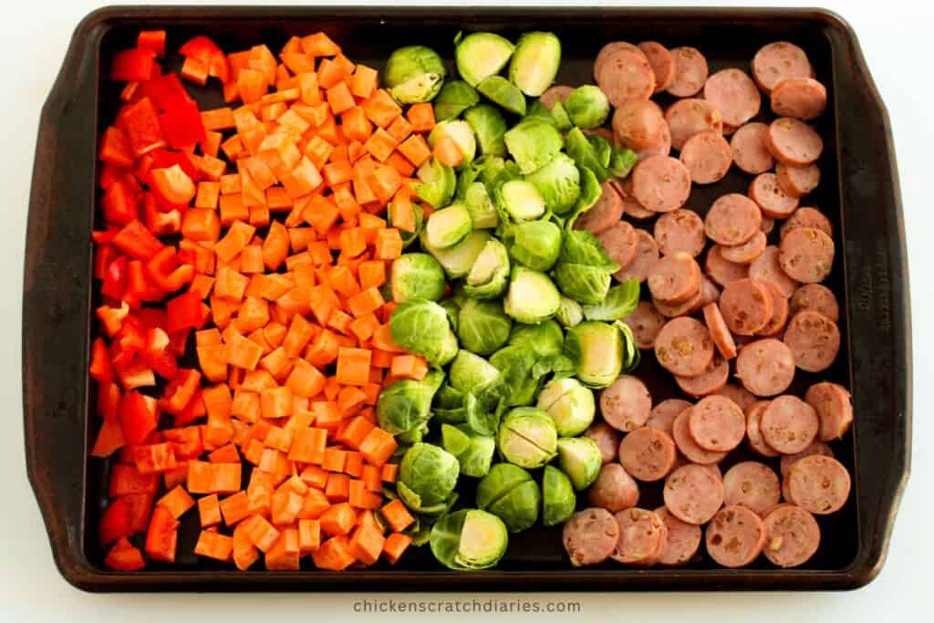 Ingredients for sheet pan meal lined up in rows on baking pan, ready to add oil and herbs mix.