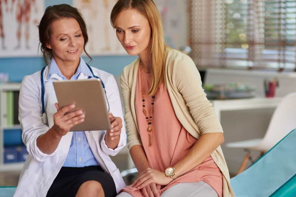 Woman sitting next to OB doctor on an exam table, looking at a tablet together.