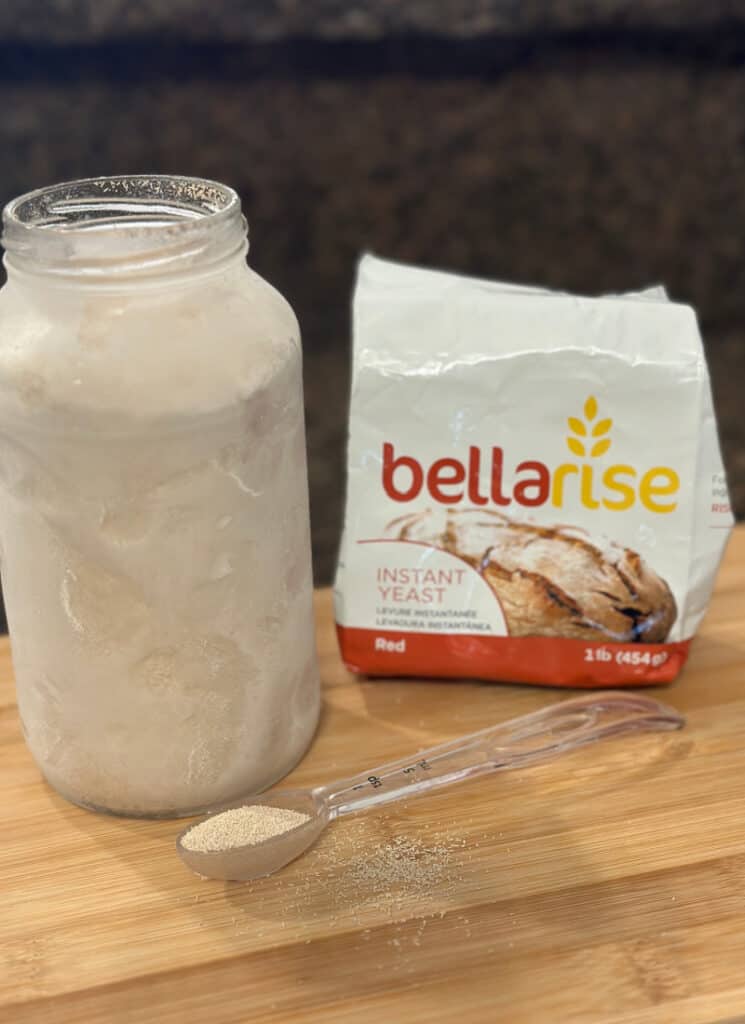 Glass Quart jar filled with yeast with a measuring spoon with yeast and a bag of yeast nearby, on countertop.