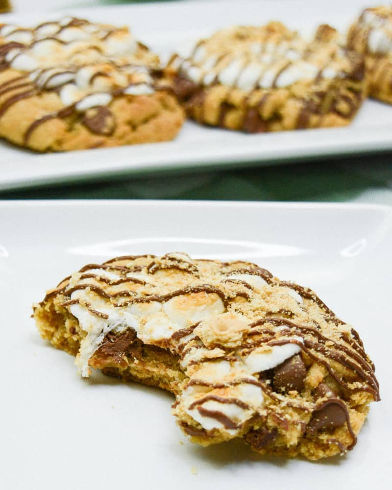 Smores cookie with a bite taken out of it on a white plate and more cookies on a tray in the background.