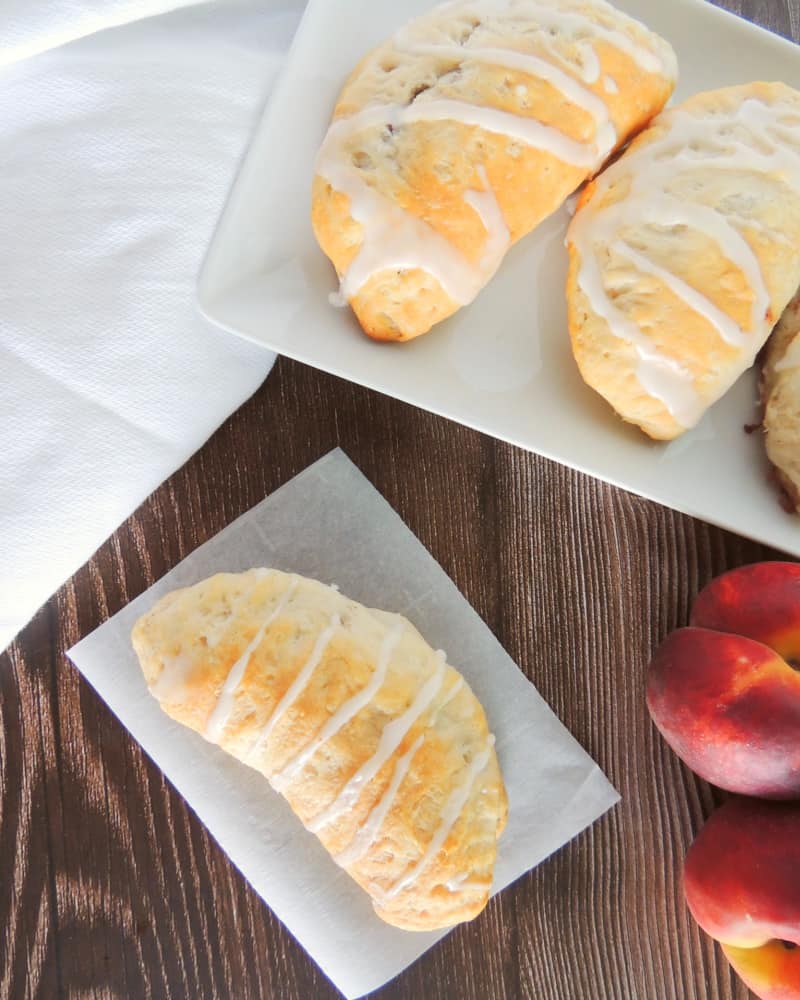 Finished glazed peach pie turnovers on a serving plate with another turnover on a piece of parchment paper nearby on a wooden table.
